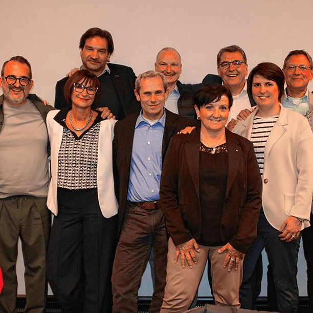 Gruppe von neun Menschen lächelt in einem semi-formalen Setting vor neutralem Hintergrund.