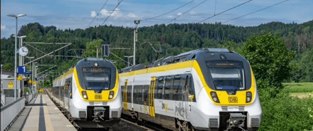 Zwei Regionalzüge der DB halten an einem Bahnhof, umgeben von einer grünen Landschaft und einem blauen Himmel.