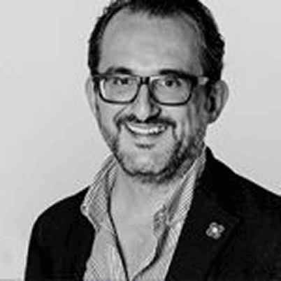 Porträt eines lächelnden Mannes mit Brille, schwarzem Sakko und kariertem Hemd, vor einfarbigem Hintergrund.