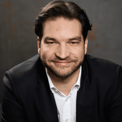 Ein lächelnder Mann mit dunklen Haaren trägt einen schwarzen Anzug und steht vor einem grauen Hintergrund.