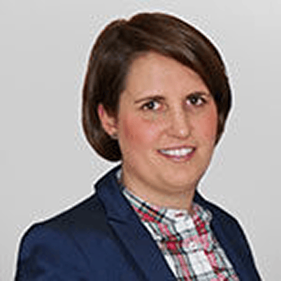 Porträt einer Person mit braunen Haaren, die einen dunkelblauen Blazer und ein kariertes Hemd trägt. Neutraler Hintergrund.