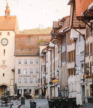 Historische Straßenansicht mit gepflegten Gebäuden, einem Uhrenturm und leeren Terrassen in sanfter Morgenbeleuchtung.
