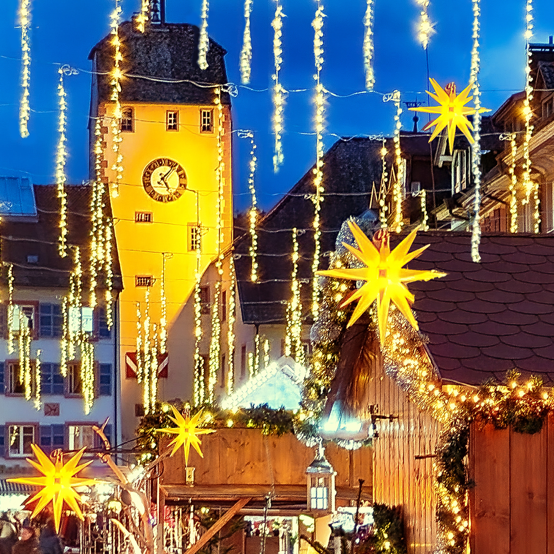 Weihnachtlicher Markt mit beleuchtetem Turm und dekorativen Sternen in einer festlichen Abendstimmung.