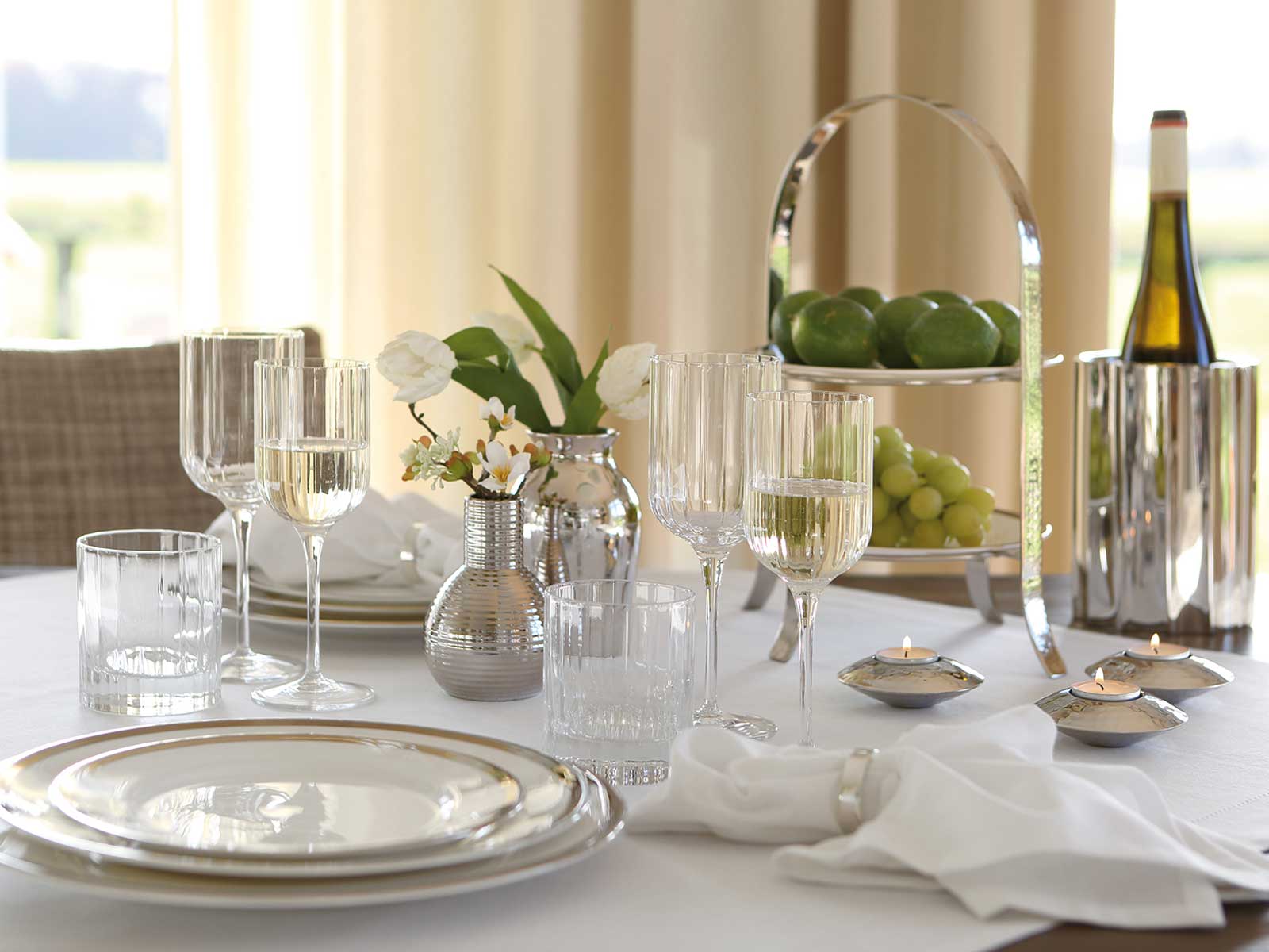 Elegant gedeckter Tisch mit Gläsern, Geschirr, einer Blumenvase, Obst und einem Weinkühler.