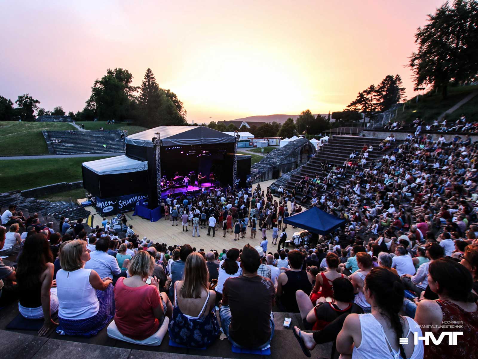Menschenmenge genießt ein Konzert in einem Amphitheater, umgeben von Natur und einer schönen Abenddämmerung.