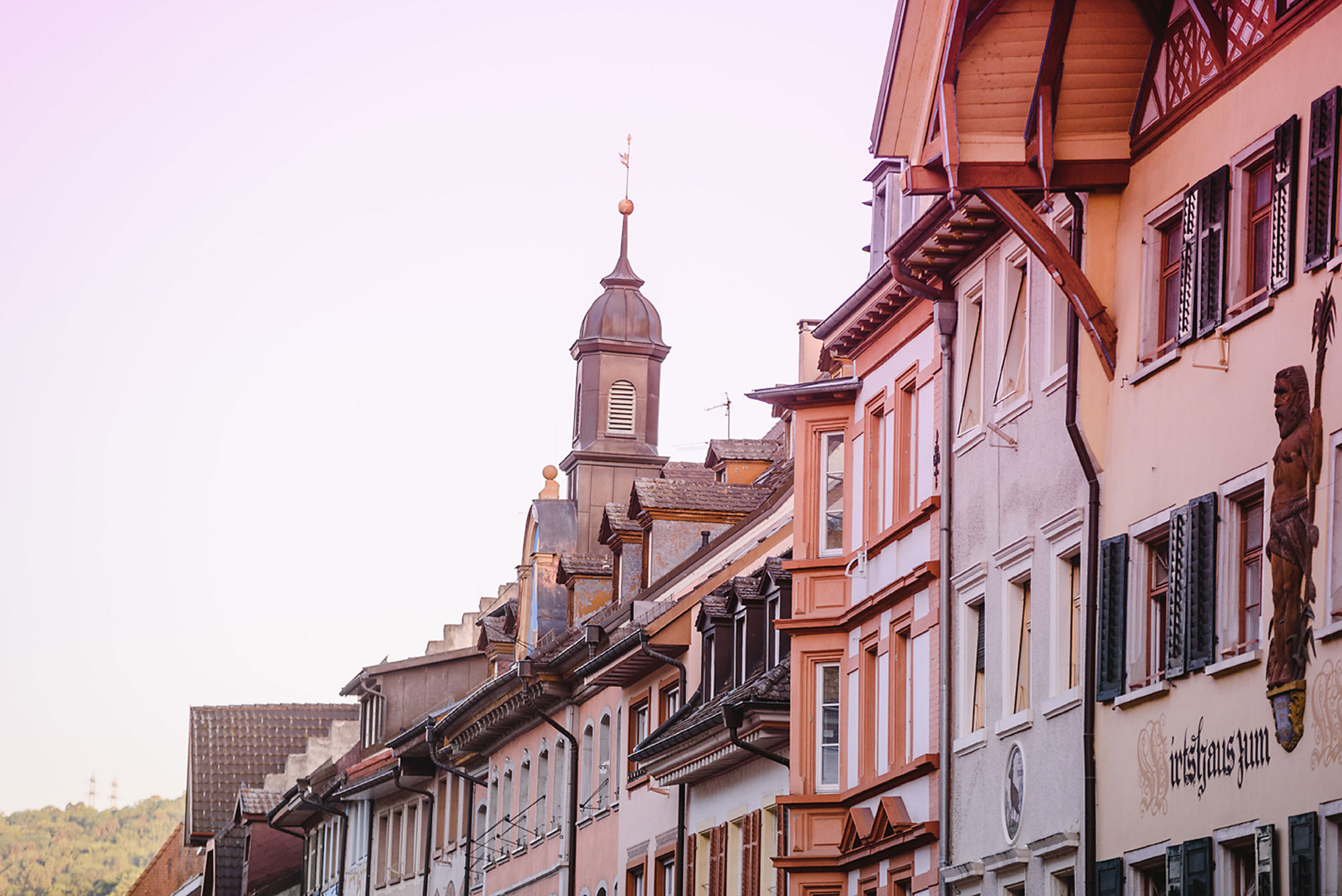 Ansicht von bunten, historischen Gebäuden mit einem Kirchturm im Hintergrund und sanften Farben am Himmel.