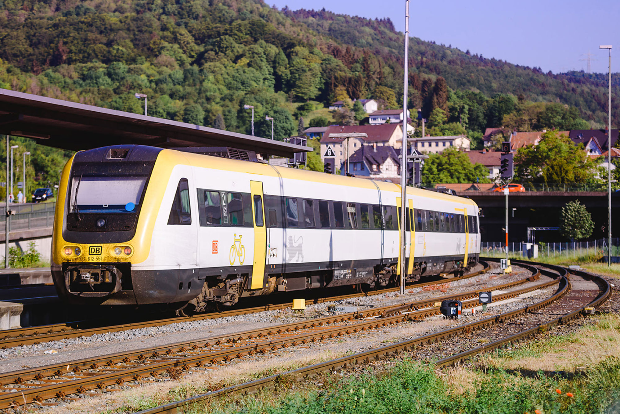 Ein gelber und grauer Triebwagen der DB auf einem Bahnhof, umgeben von grünen Hügeln und Häusern.