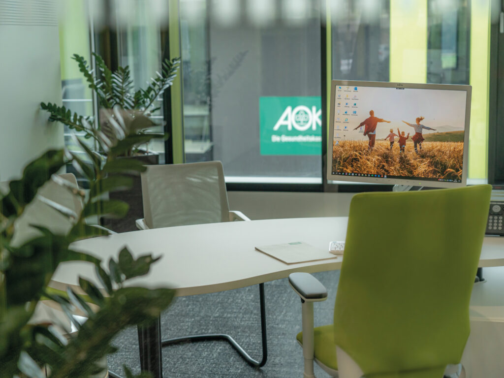 Moderne Büroansicht mit Schreibtisch, Computer und Pflanzen im Vordergrund, Fenster mit Blick auf AOK-Logo.
