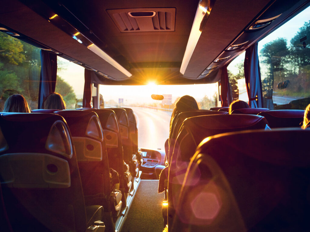 Blick aus einem Reisebus auf die Straße, mit Sonnenaufgang im Hintergrund und Passagieren in den Sitzen.