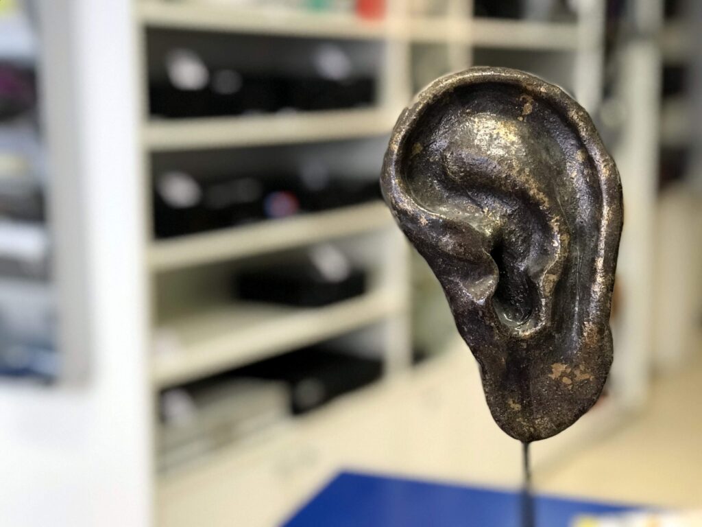 Eine bronzene Skulptur eines menschlichen Ohrs steht auf einem Tisch. Im Hintergrund sind Regale zu erkennen.