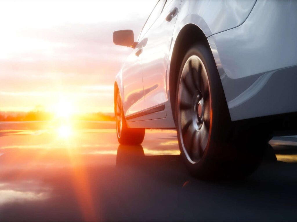 Nahaufnahme eines weißen Autos mit Sonnenuntergang im Hintergrund, das auf einer nassen Straße fährt.