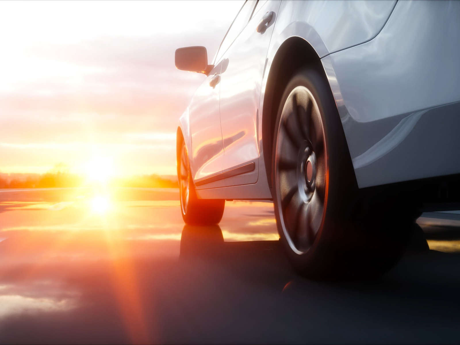 Nahaufnahme eines weißen Autos mit Sonnenuntergang im Hintergrund, das auf einer nassen Straße fährt.