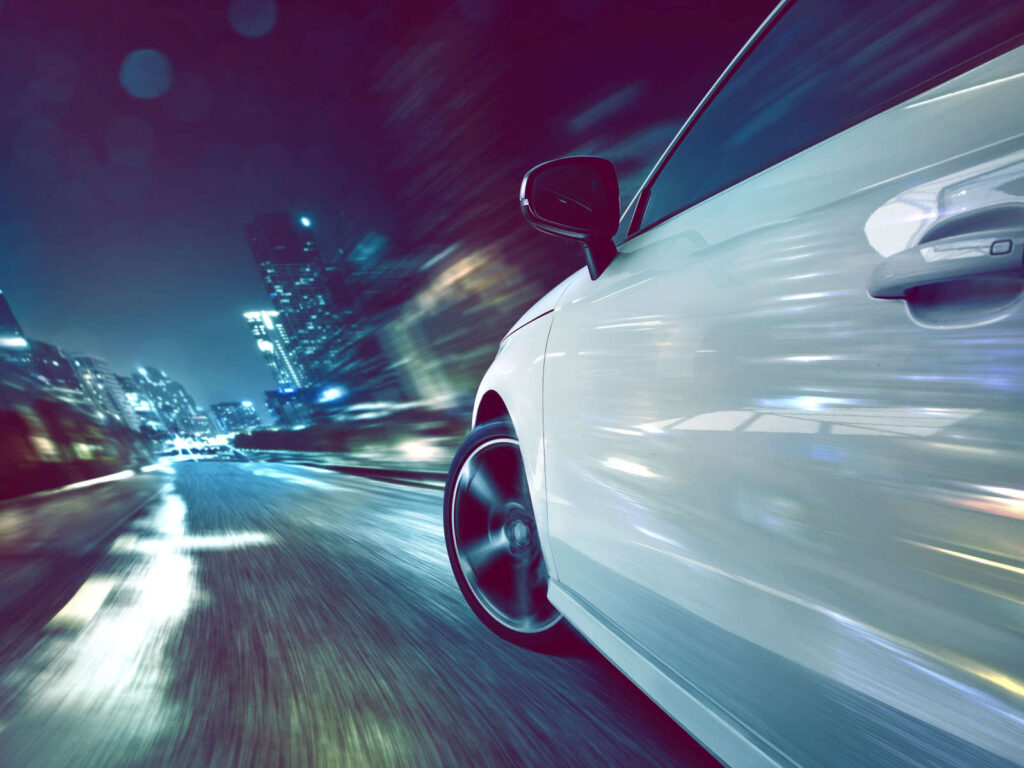 Ein Weißes Auto fährt schnell auf einer beleuchteten Stadtstraße bei Nacht, lichtspuren im Hintergrund.