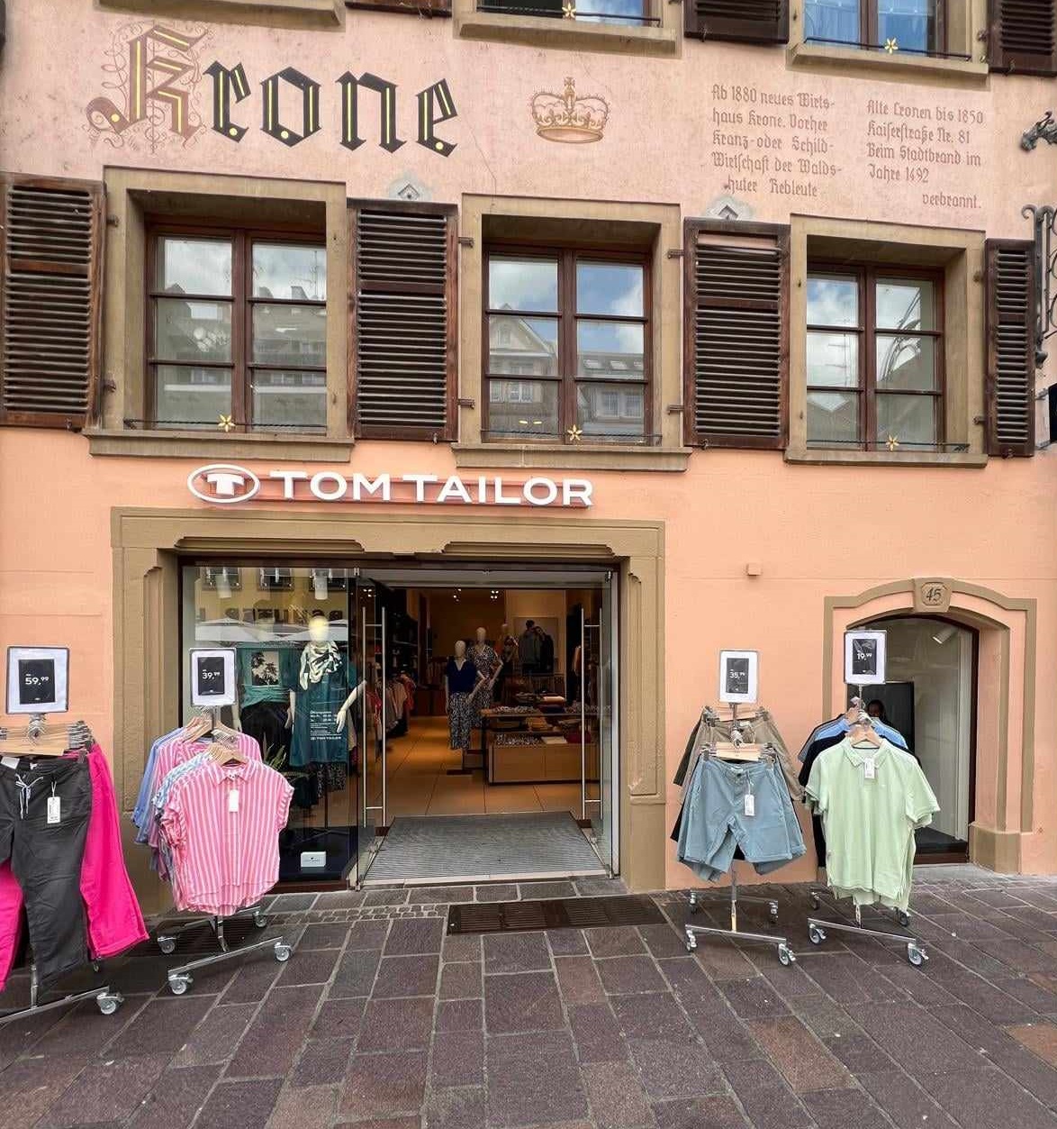 Schaufenster eines Tom Tailor-Geschäfts mit Kleidung und einer Fassade, die den Schriftzug "Krone" zeigt.