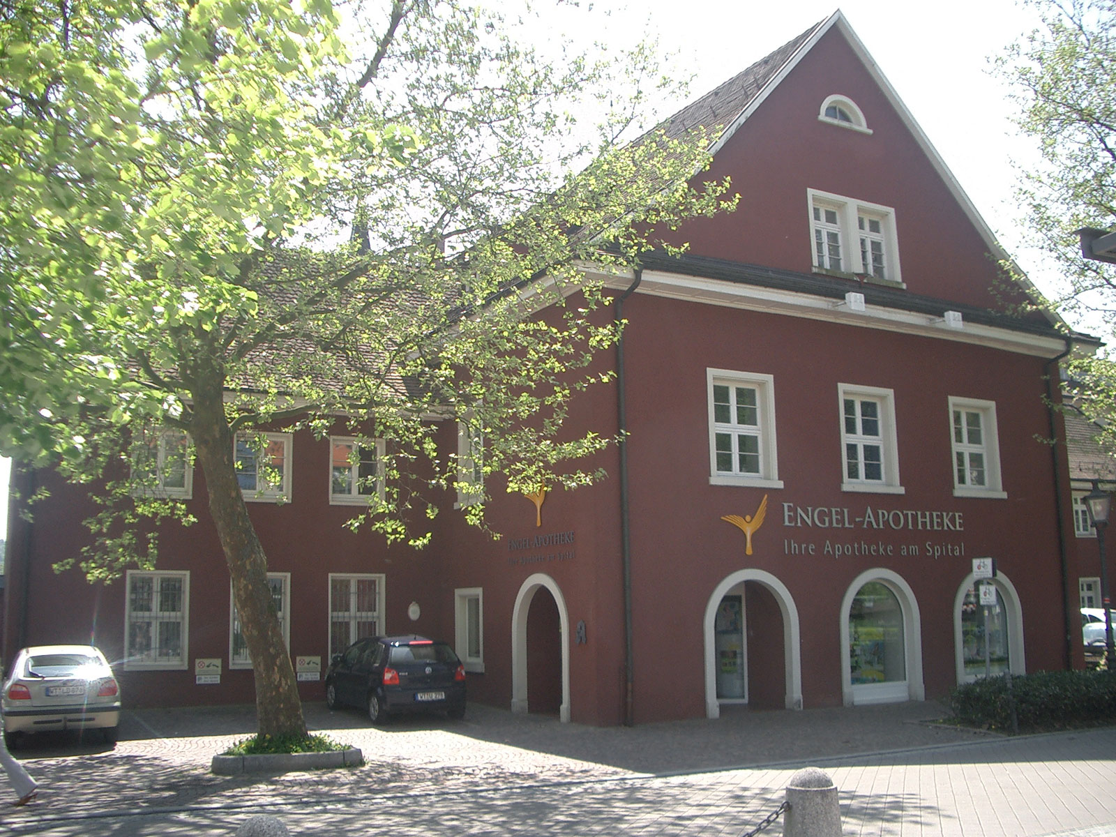 Rote Apotheke namens "Engel-Apotheke" mit Blick auf belebten Platz, umgeben von Bäumen und Pkw.