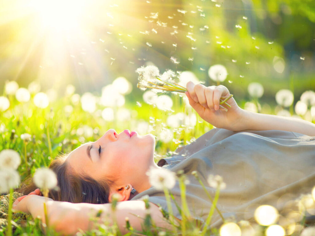 Junge Frau liegt auf Wiese, bläst Pusteblumen und genießt die Sonne, umgeben von schimmernden Dandelion-Samen.