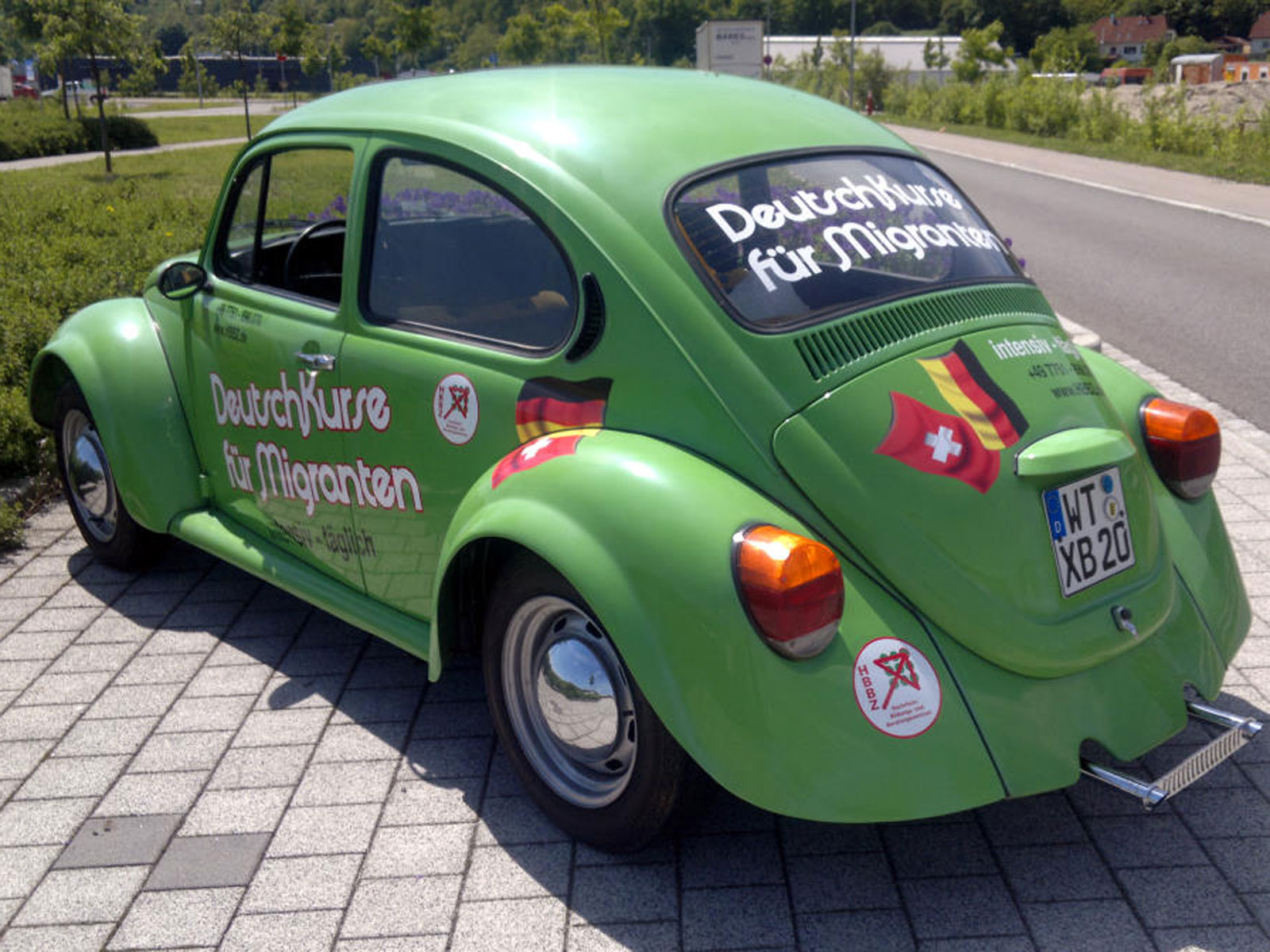 Grüner Volkswagen Käfer mit Aufschrift „Deutschkurse für Migranten“ steht auf einem Parkplatz.