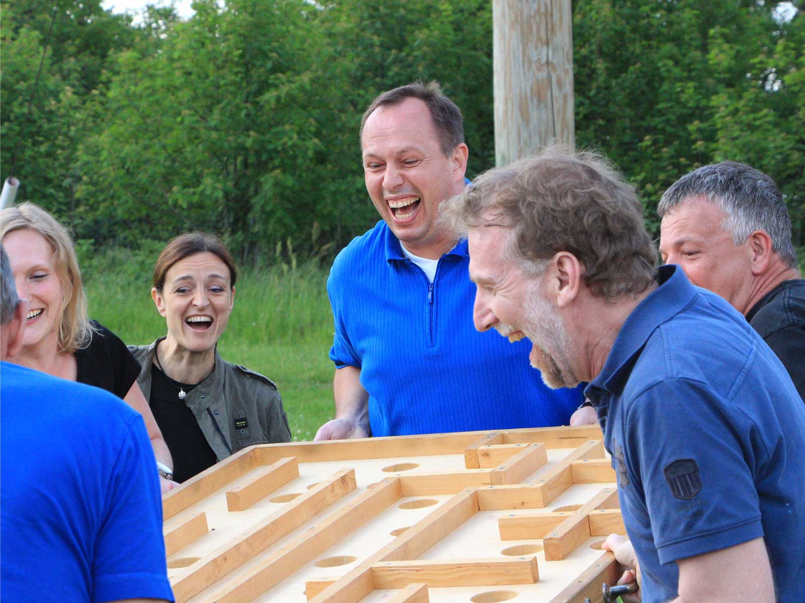 Eine Gruppe von Menschen lacht und hebt gemeinsam ein Holzspiel, umgeben von grüner Wiese.