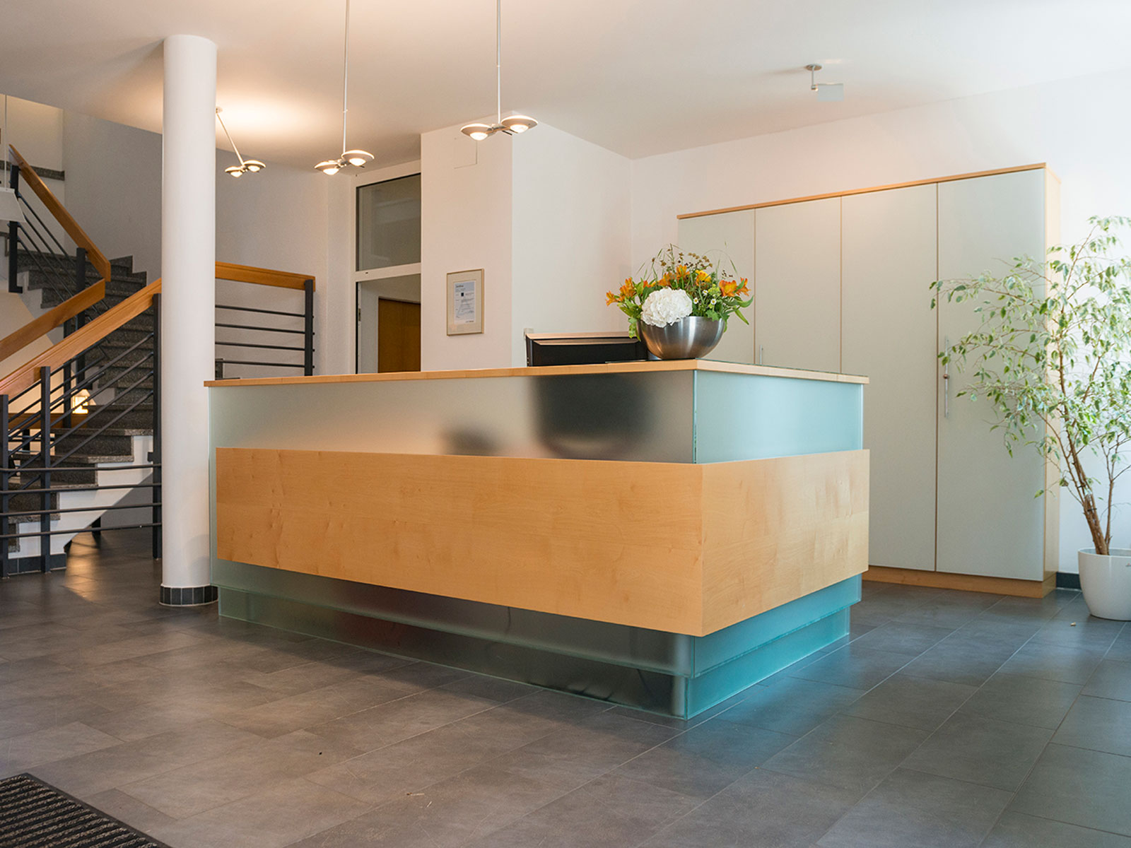 Moderne Empfangstheke mit Holz- und Glaselementen in hellen Räumen, umgeben von Pflanzen und Treppenaufgang.