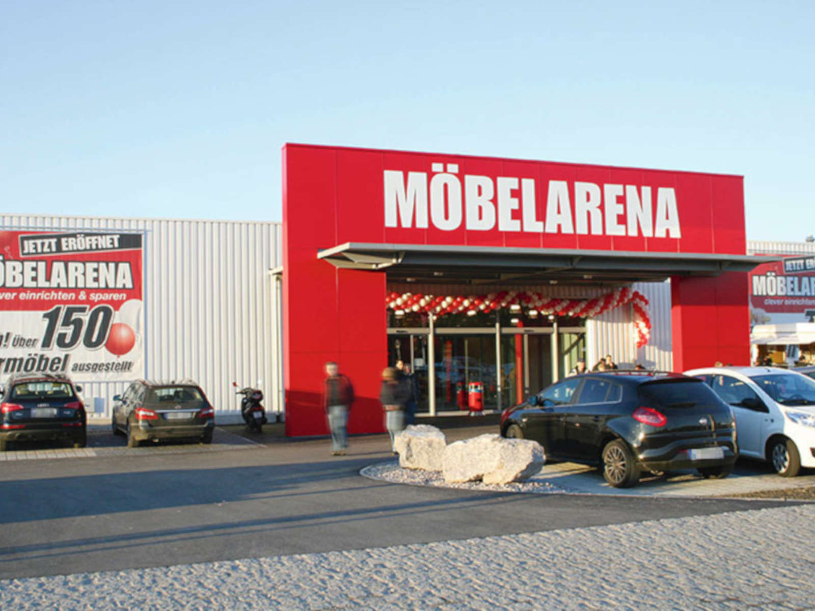 Eingang zur Möbelarena mit Autos im Parkbereich und Werbebanner zur Eröffnung und Möbelangebot.