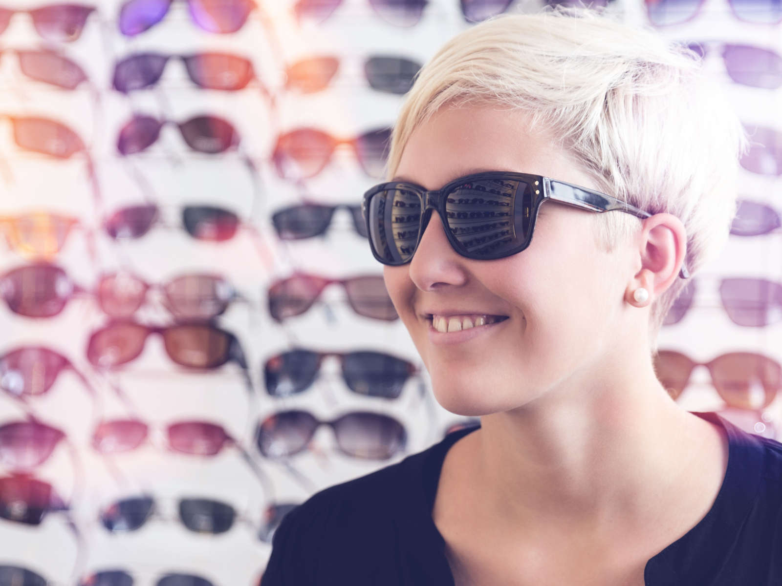 Eine junge Frau mit kurzen, blonden Haaren und Sonnenbrille lächelt vor einer Wand mit verschiedenen Sonnenbrillen.