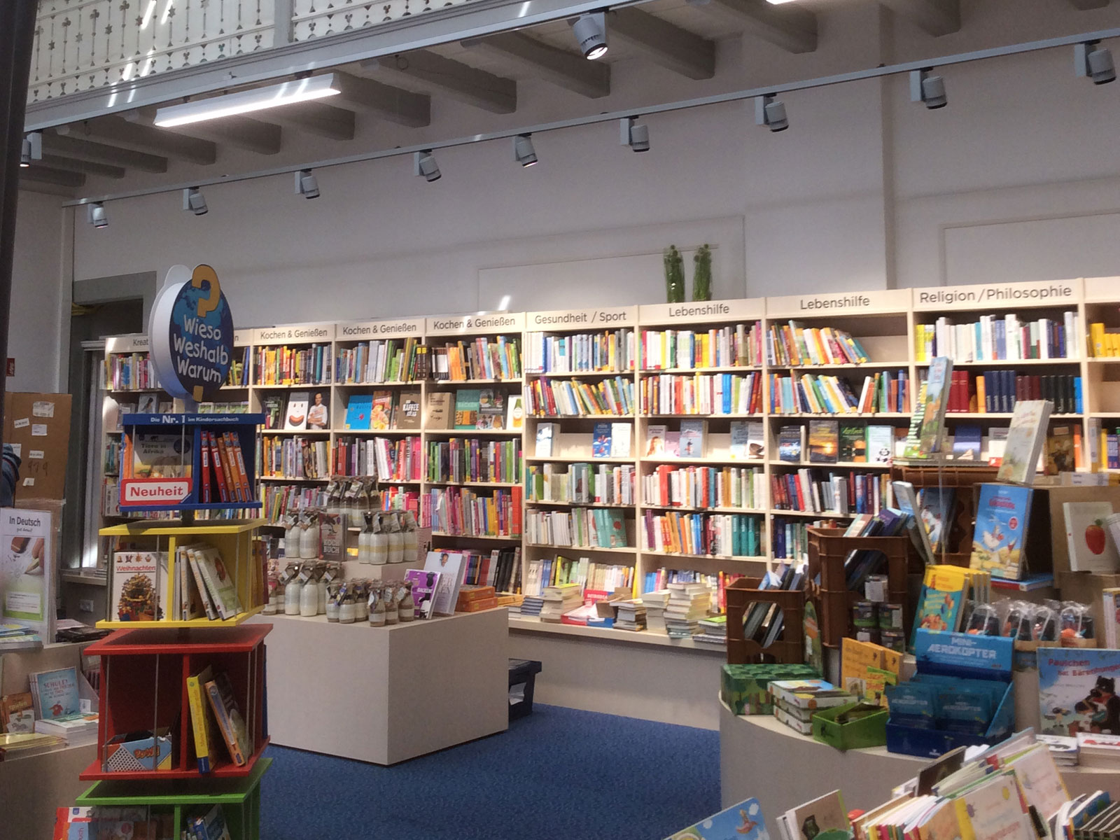 Blick in eine Buchhandlung mit Regalen voller Bücher und einem bunten Tisch mit Neuheiten und Geschenken.