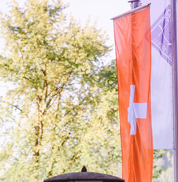 Zwei Fahnen wehen im Wind, eine orangerot mit einem weißen Kreuz, die andere mit einem Symbol, vor einem Baum.