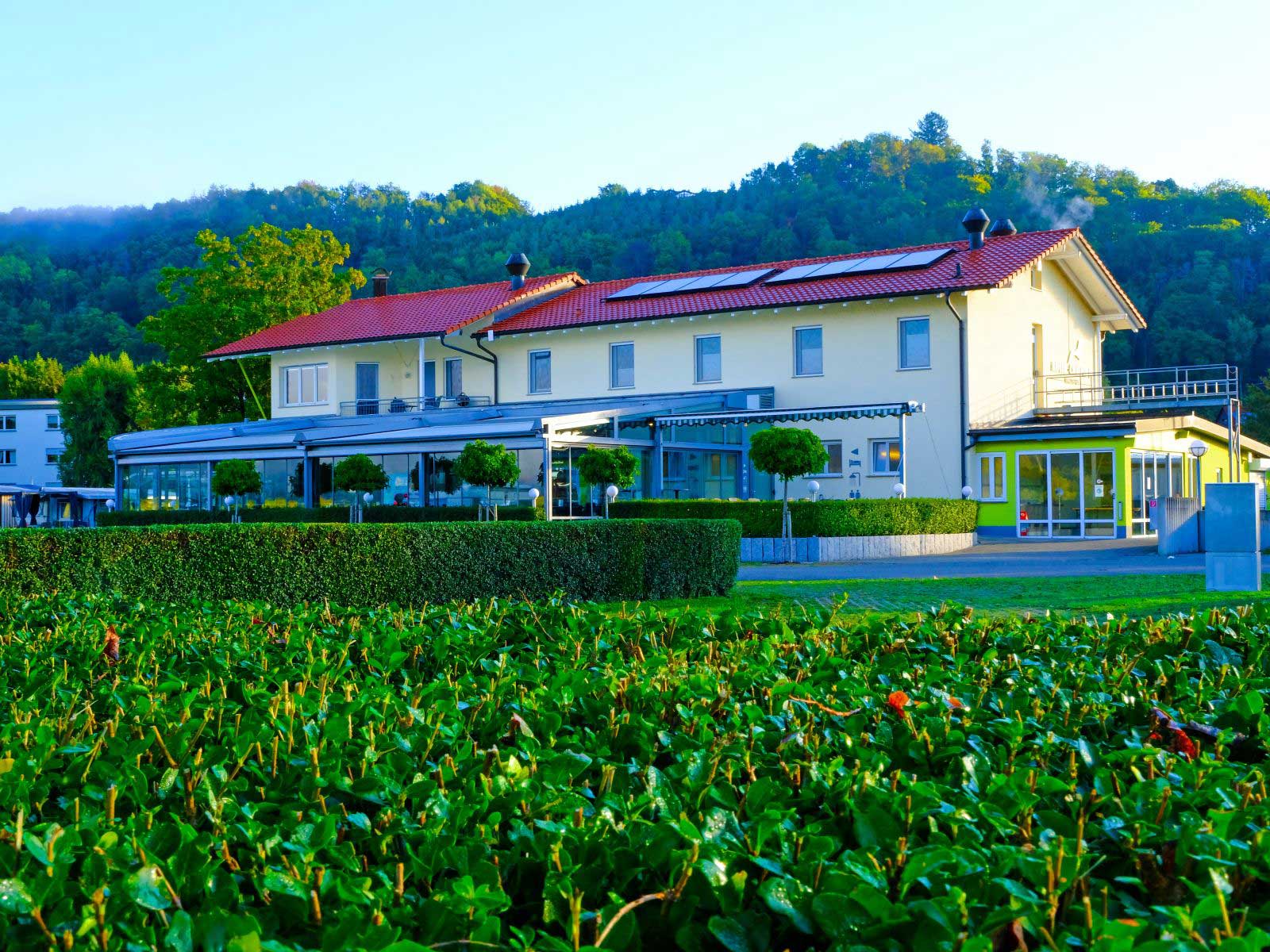 Modernes Gebäude mit rotem Dach, umgeben von Hecken und Bäumen, gelegen in einer grünen Landschaft unter blauem Himmel.