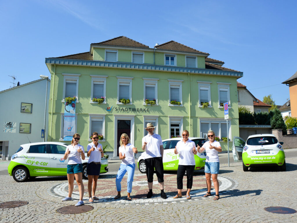 Sechs Personen in weißen T-Shirts posieren vor einem grünen Stadthaus und zwei grünen Autos auf einem Platz.