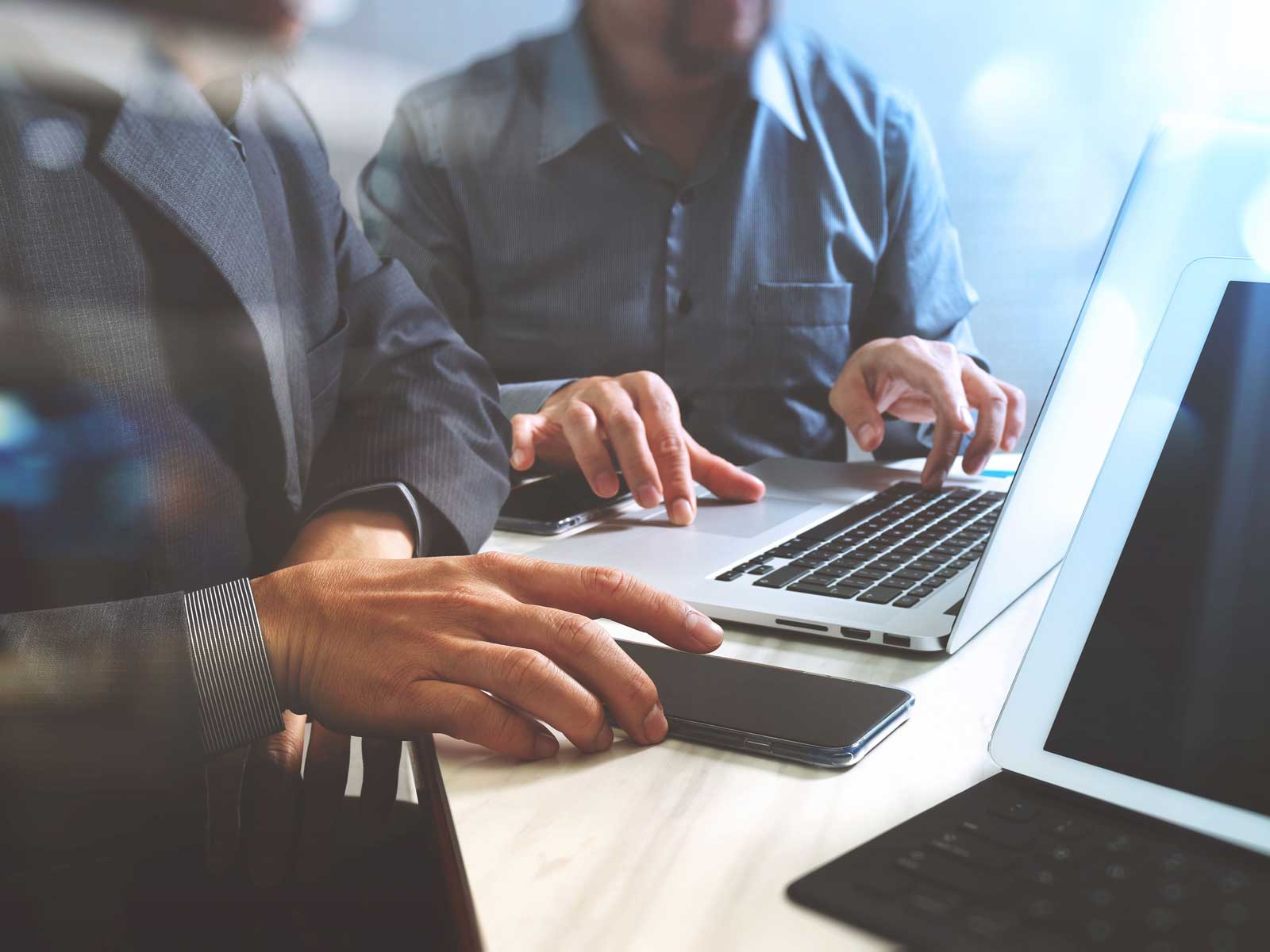 Zwei Personen in Anzügen nutzen Laptops und ein Smartphone, während sie an einem Tisch arbeiten.