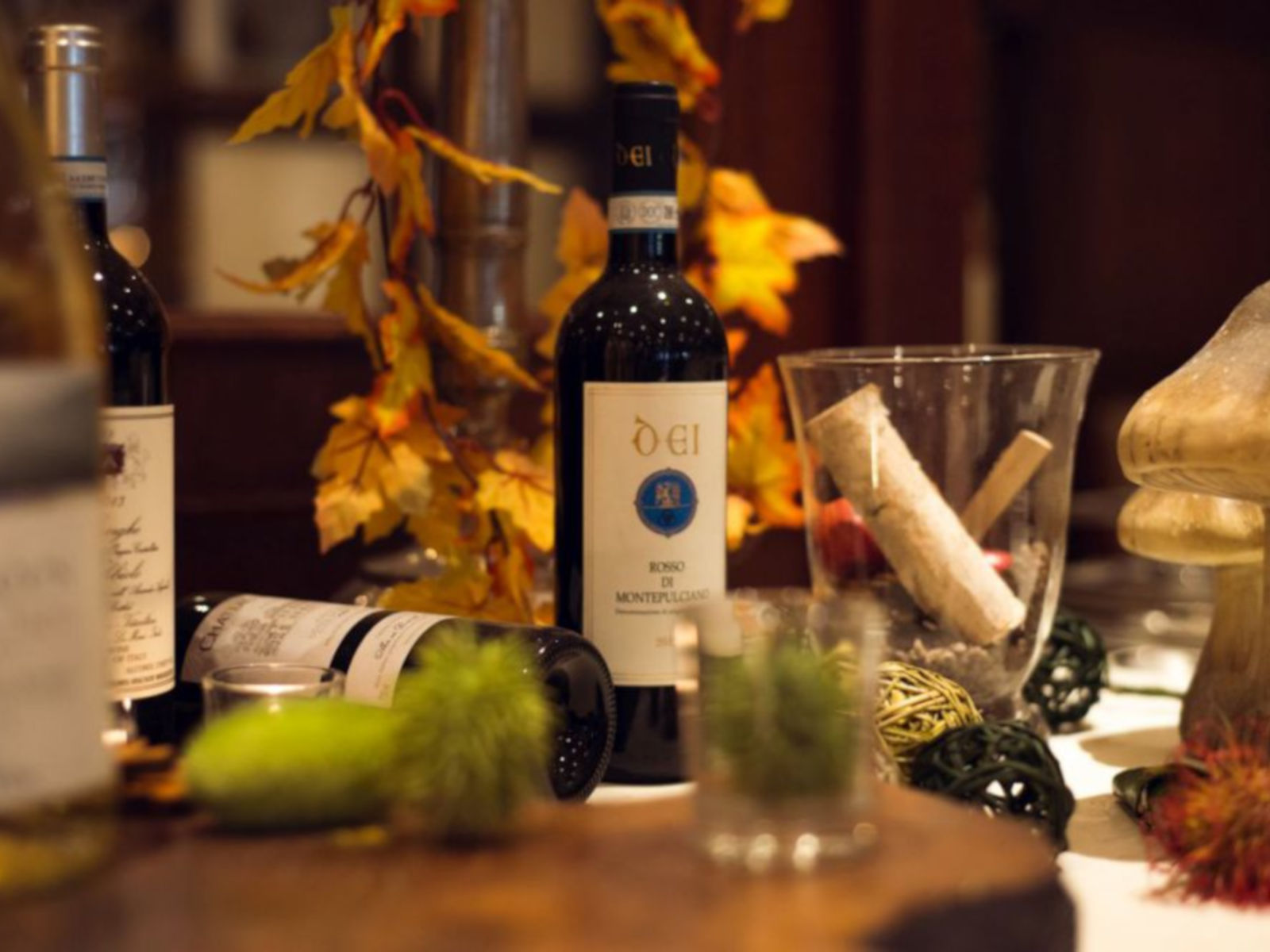 Stillleben mit Weinflaschen, Gläsern, dekorativen Elementen und buntem Herbstlaub auf einem Tisch.