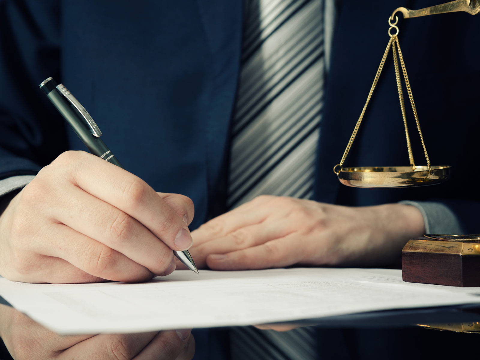 Eine Hand mit einem Stift schreibt auf Dokumenten, während eine Justitia-Waage im Hintergrund steht.