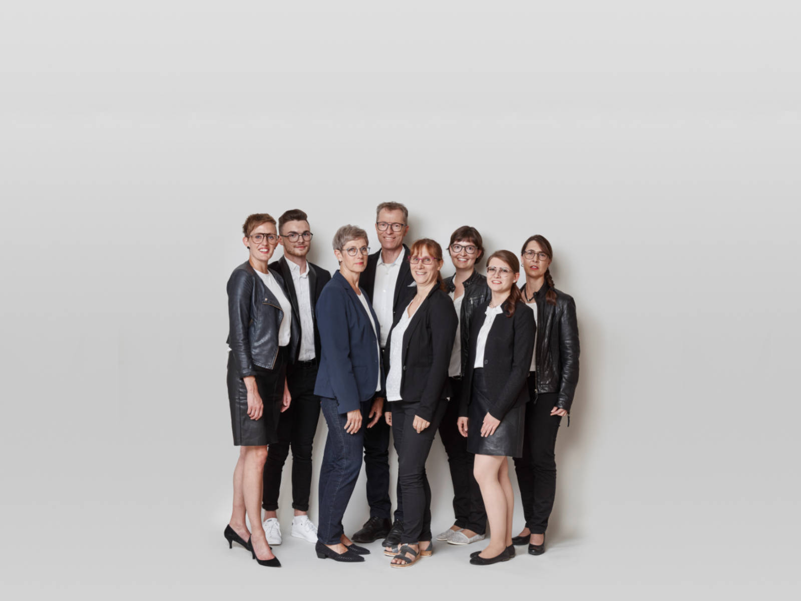 Eine Gruppe von neun Personen in formeller Kleidung steht vor einem hellen Hintergrund.