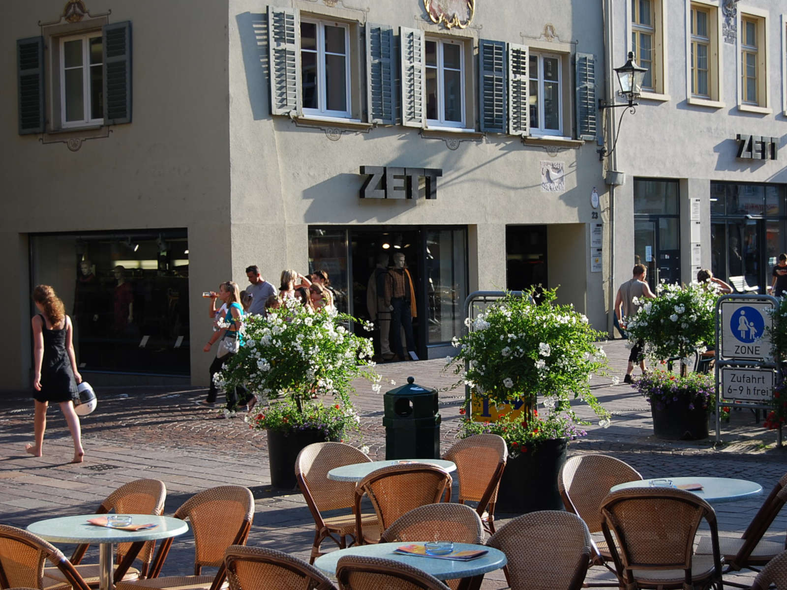 Ein belebter Platz mit Cafétischen, Blumen und Passanten vor einem Bekleidungsgeschäft.