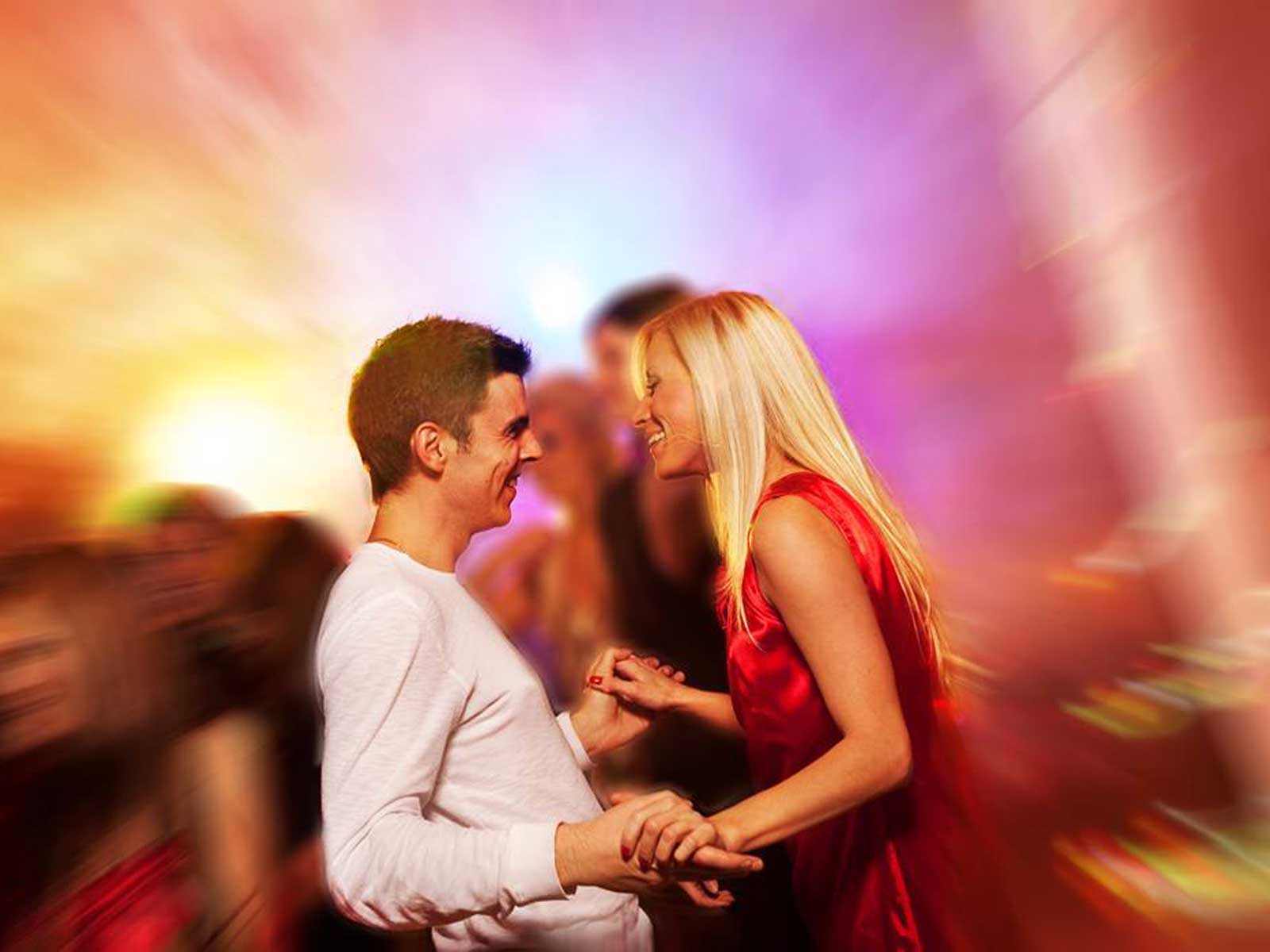 Ein Paar tanzt lebhaft in einer bunten, verschwommenen Partyszene. Sie lächeln und genießen den Moment.