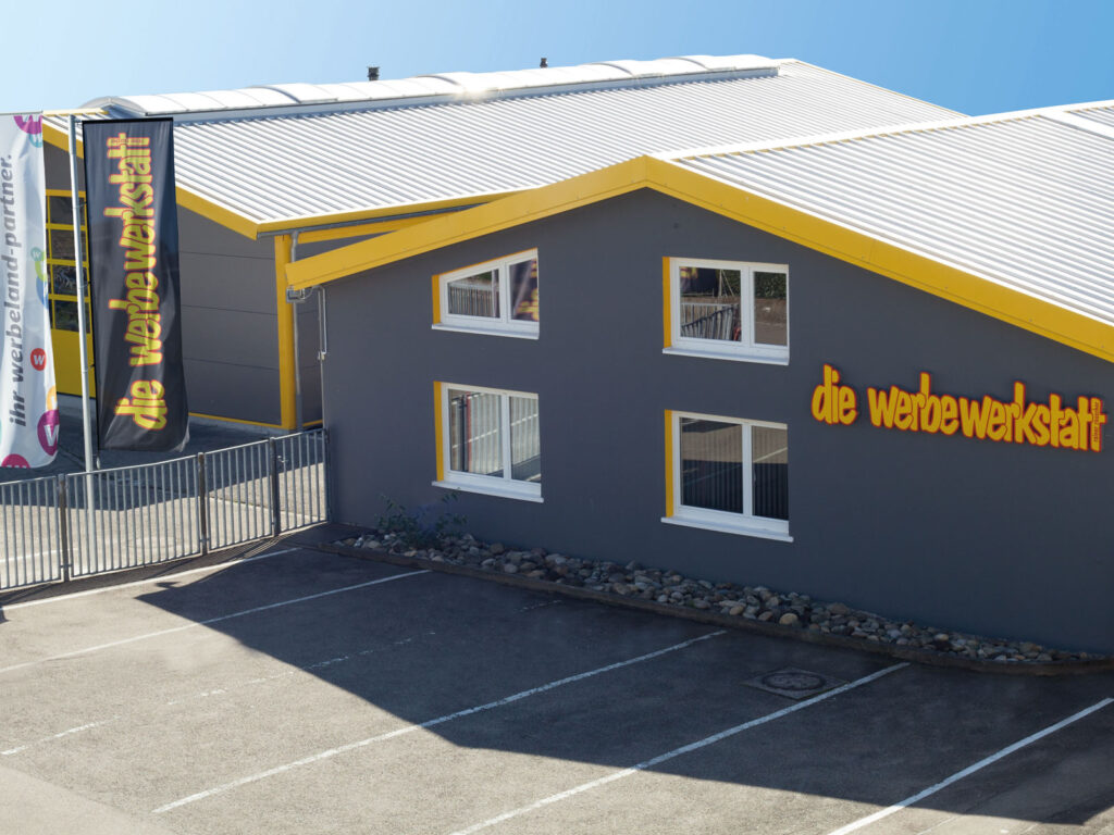 Fassade eines grauen Gebäudes mit gelben Akzenten und dem Schriftzug „die werbewerkstatt“. Parkplatz im Vordergrund.