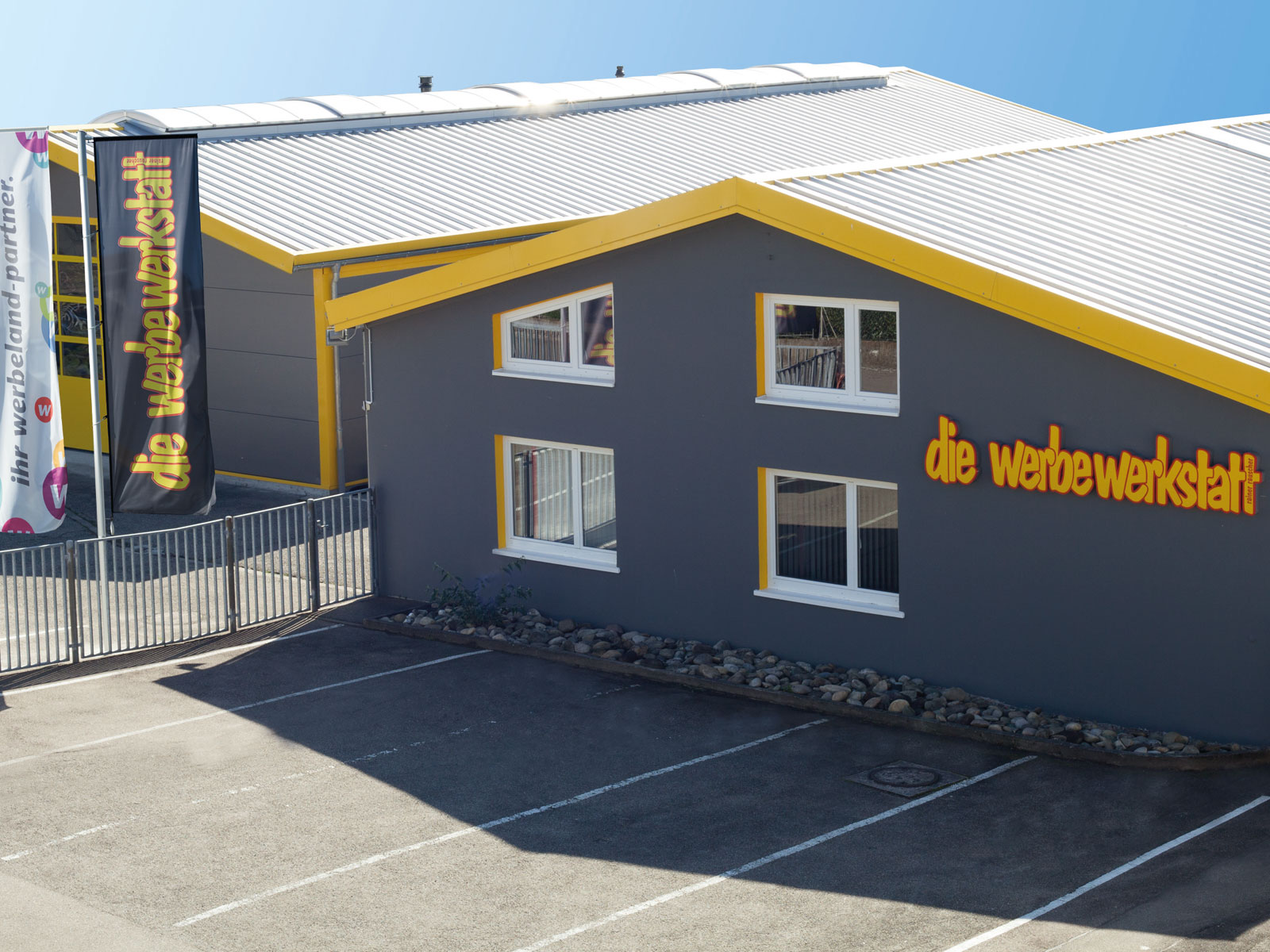 Fassade eines grauen Gebäudes mit gelben Akzenten und dem Schriftzug „die werbewerkstatt“. Parkplatz im Vordergrund.