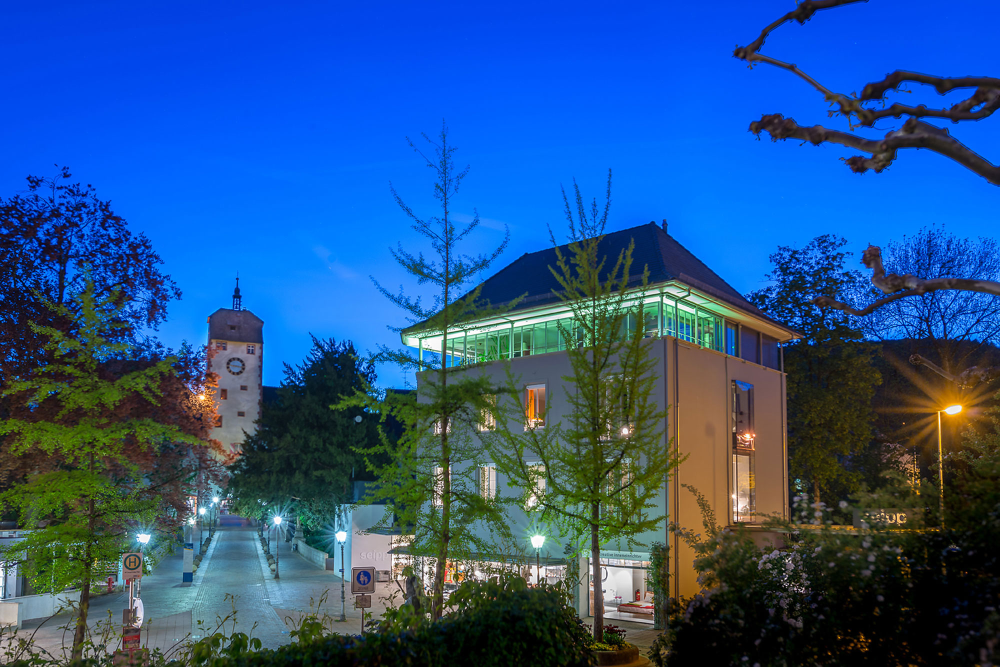 Stadtansicht bei Abenddämmerung mit modernem Gebäude und einem alten Uhrturm im Hintergrund.