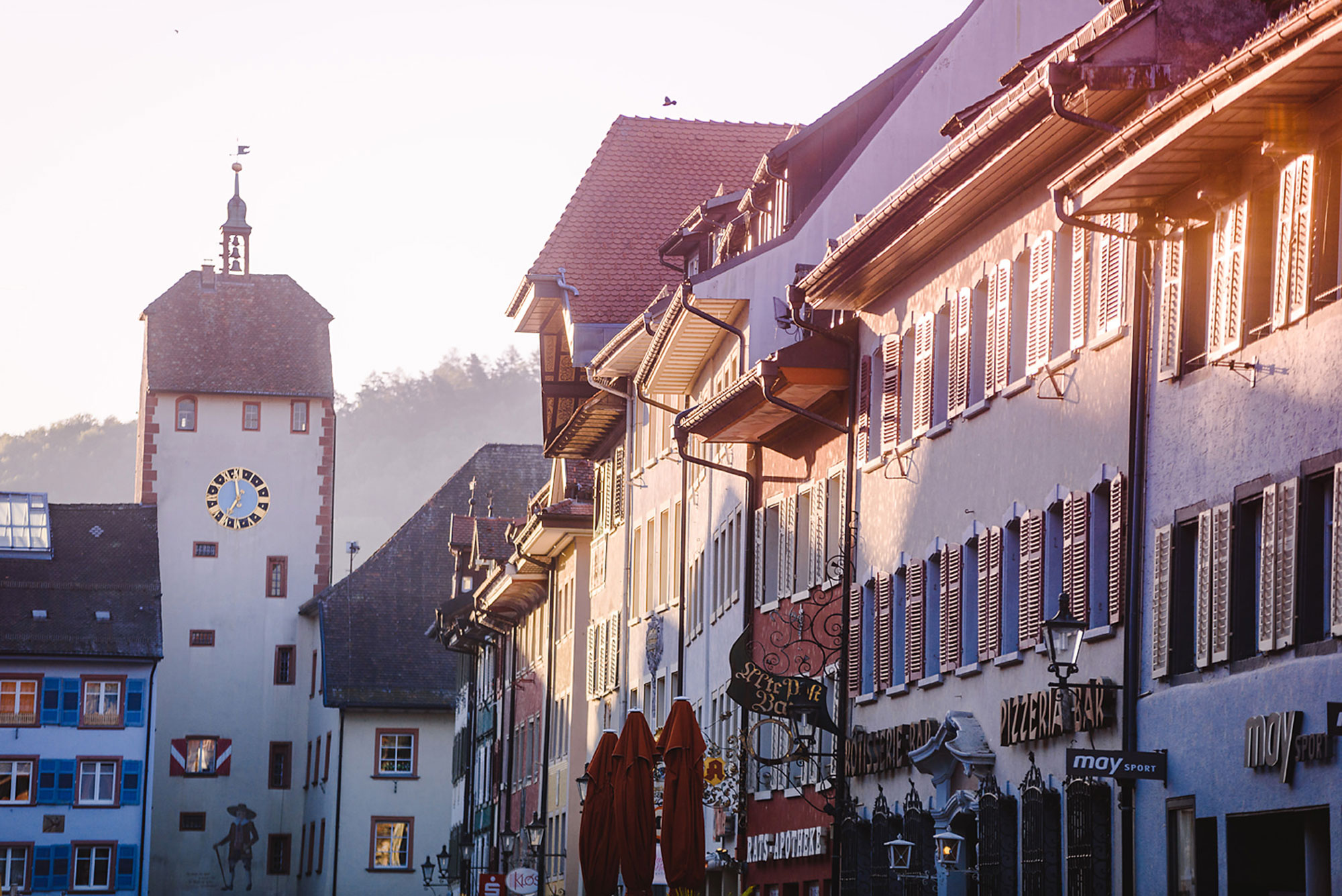 Sonnenlicht beleuchtet historische Gebäude mit einem Uhrturm in einer malerischen Stadtstraße.
