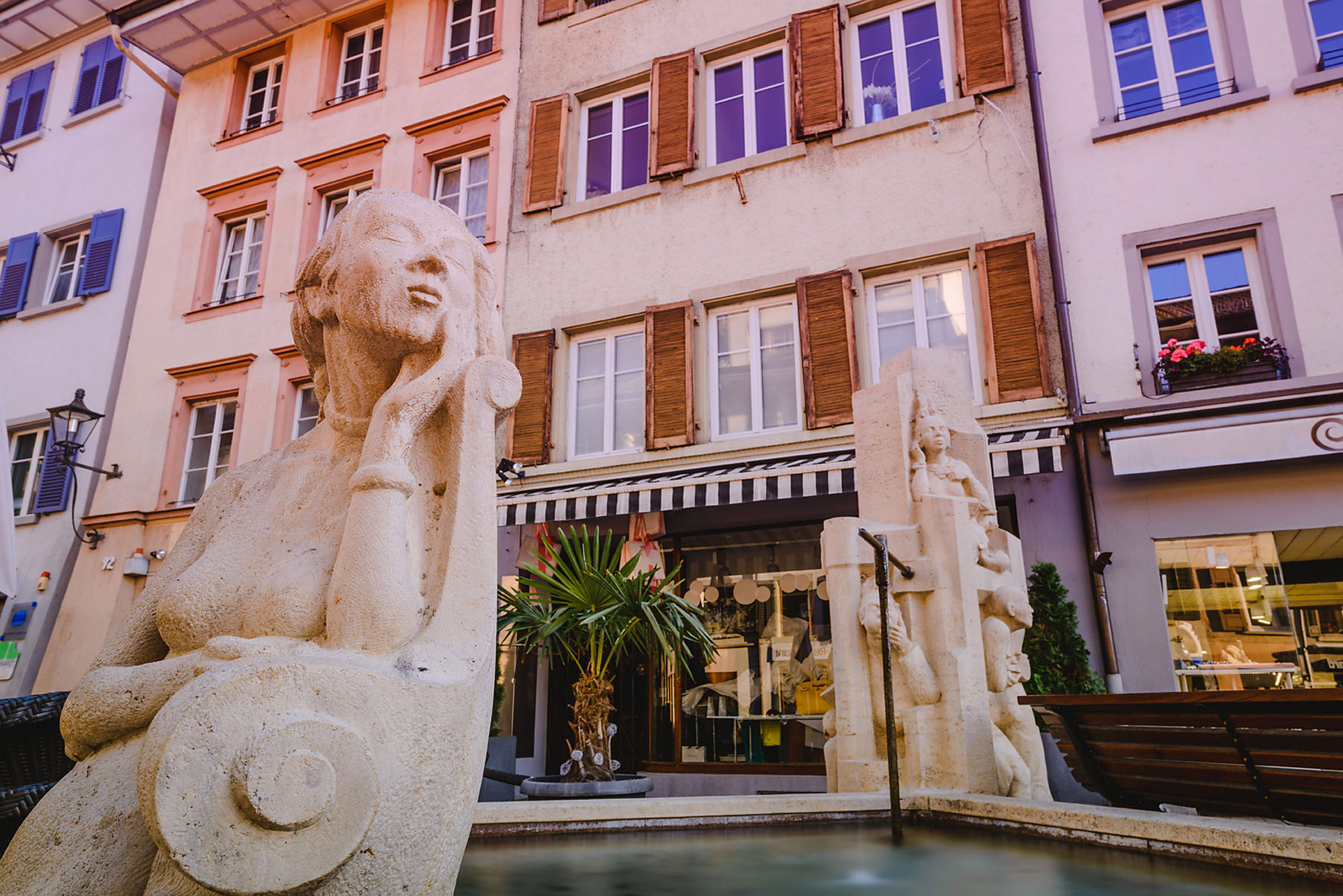 Skulpturen in einem Platz, umgeben von historischen Gebäuden und einem kleinen Wasserbecken.