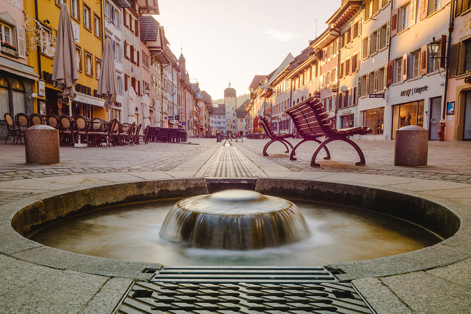 Ruhige Stadtstraße mit einem Wasserspielelement und Sitzbänken, umgeben von bunten Gebäuden.