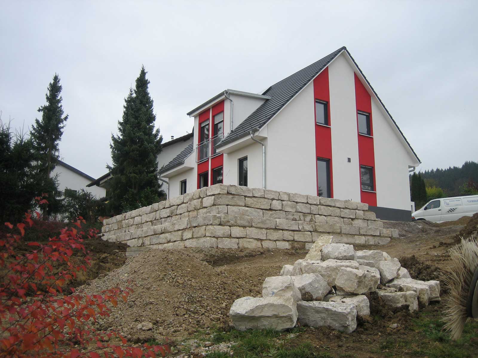 Moderner, zweistöckiger Wohnhaus mit roter Akzentwand und Steinterrasse im Vordergrund, umgeben von Natur.
