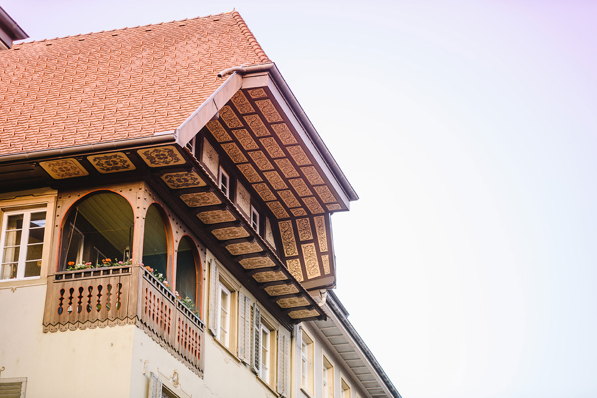 Detailansicht eines historischen Gebäudes mit einem dekorativen Dachvorsprung und blühenden Pflanzen auf dem Balkon.