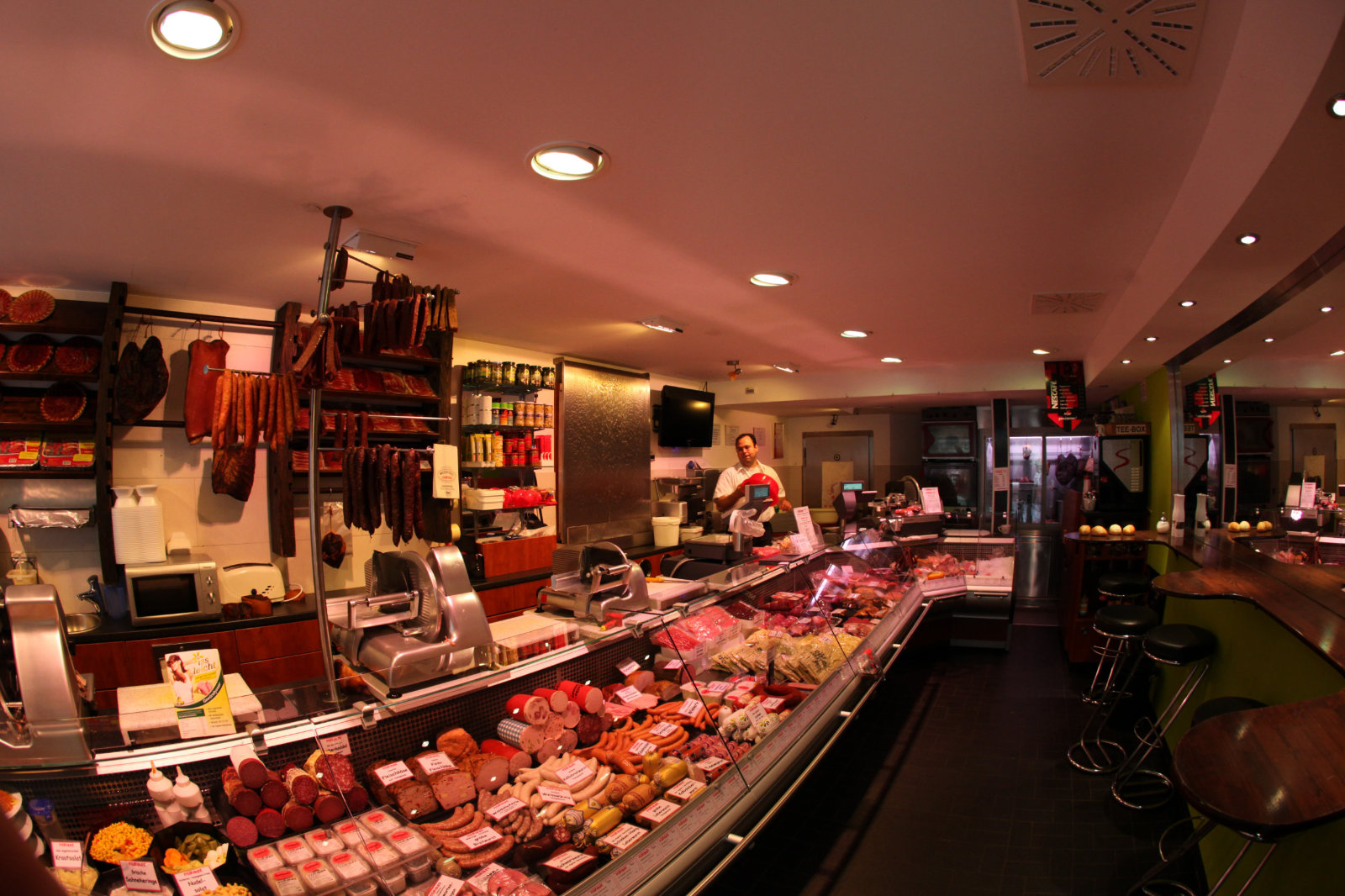 Metzgerei mit einer Vielzahl an Fleischwaren, Theken und einem Verkäufer im Hintergrund. Moderne Einrichtung und Beleucht...