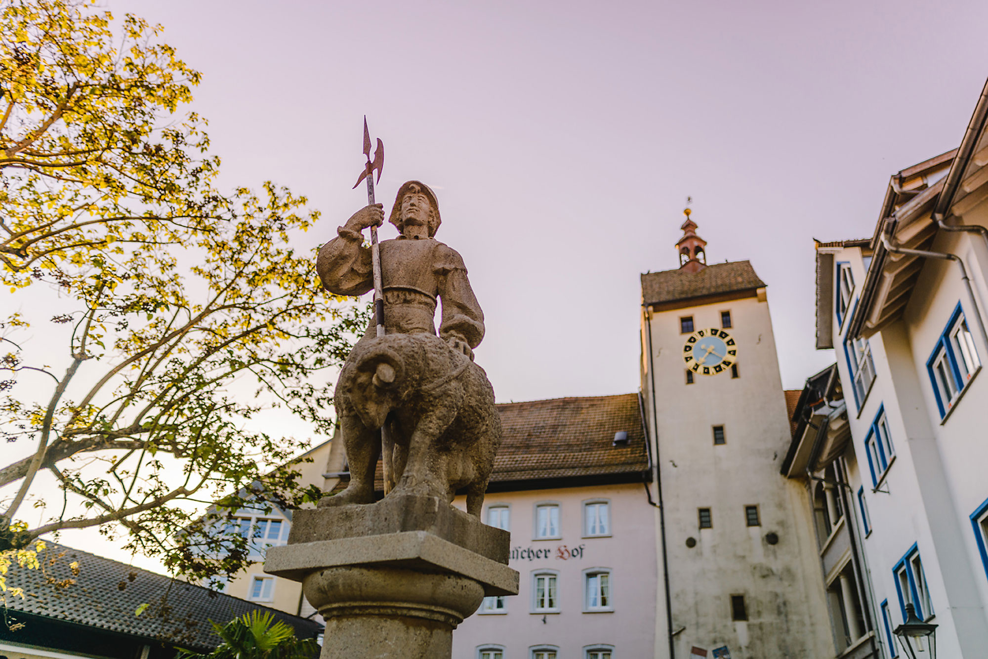 Skulptur eines Kriegers mit Lanze, der auf einem Tier steht, im Vordergrund eines historischen Gebäudes mit Uhr.