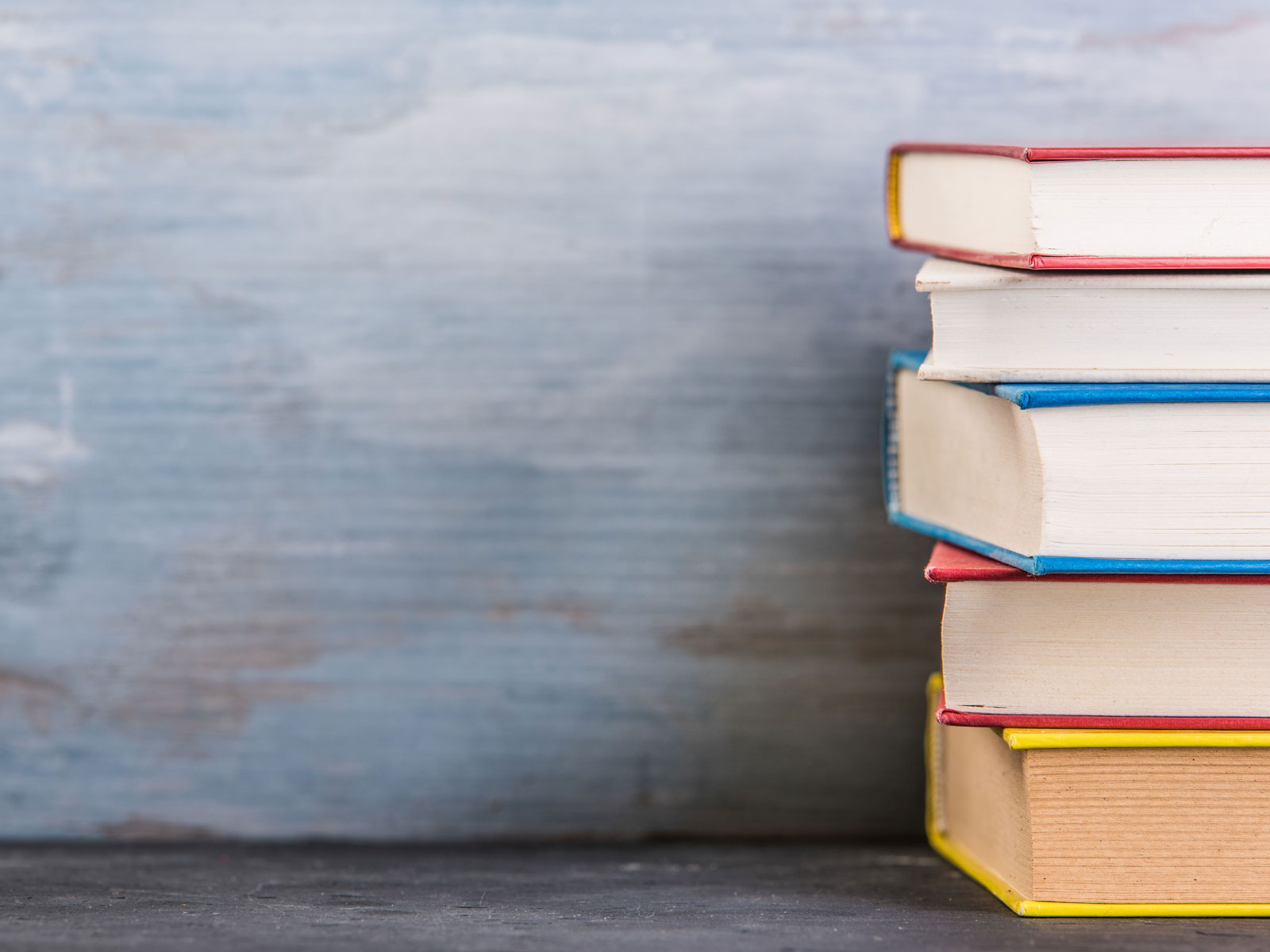 Ein Stapel Bücher mit bunten Buchrücken steht auf einer dunklen Oberfläche vor einem blauen Hintergrund.